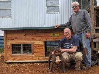 Freedom For Fido crew leader Fritz Gilbert, left, and volunteer Tim Pauley stand with Ally and her new doghouse during a recent fence build. Pauley custom built the doghouse for Ally as she and her family received a free fence from the non-profit group. More doghouses are needed for future builds and there is a way the community can help and possibly win a contest as well.