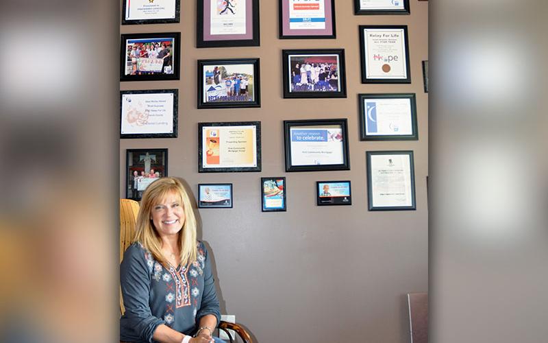 Tracie Griffith, owner and mortgage broker of First Community Mortgage, sits next to the office’s wall showcasing many of the causes the business has sponsored and supported over the years, such as Relay for Life, St. Jude and Toys for Tots.