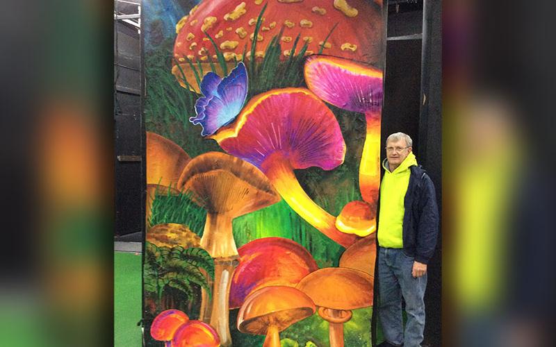 Enjoy the hand-painted murals by local artists while being entertained by the cast’s performance of Alice in Wonderland. The play began January 24 and will continue through February 9. Shown is Larry Minor standing next his mural of fantastical mushrooms.