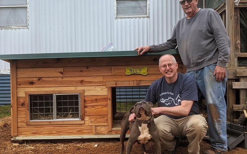Freedom For Fido crew leader Fritz Gilbert, left, and volunteer Tim Pauley stand with Ally and her new doghouse during a recent fence build. Pauley custom built the doghouse for Ally as she and her family received a free fence from the non-profit group. More doghouses are needed for future builds and there is a way the community can help and possibly win a contest as well.
