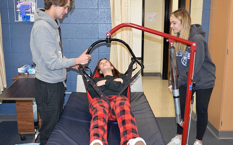 Certified Nursing Assistant (CNA) students Michael Treon and Riley Reebes demonstrate the proper way to use a life for getting a patient out of a bed using student Devan Hogsed as the patient.