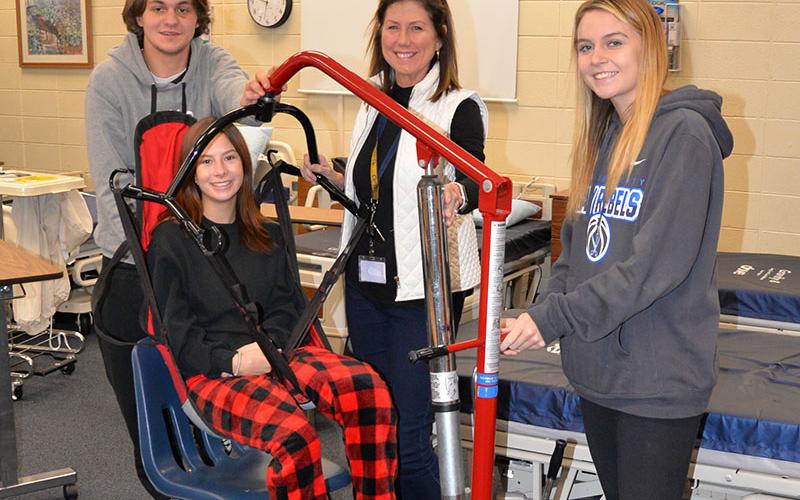 Certified Nursing Assistant (CNA) students at Fannin County High School learn to use a patient lift to move patients from a bed to a chair as part of their training. Demonstrating the equipment are, from left, Michael Treon, Devan Hogsed (seated), instructor Anne Gibbs and Riley Reeves.