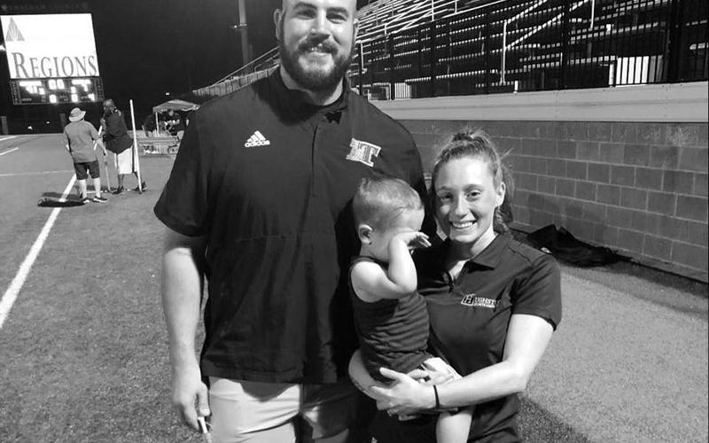 Logan Daves is pictured smiling with his wife Macy and son Laiken on the field at Toombs County High School where Daves serves as the Offensive Coordinator and assistant coach of the wrestling team.