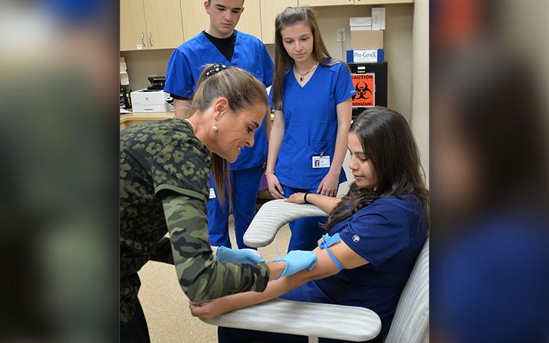 Fannin County High School juniors Trent Davis and Blair Deal are shown by Candy Hill how to prep a patient to draw blood. The example patient is Stephanie Torres, a staff member at Georgia Mountains Health. This was an exercise for the general medicine students in their clinical rotation at the facility.
