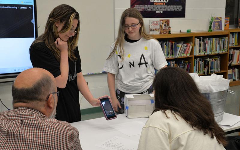 First place winners of Fannin County Middle School’s Shark Tank, Caroline Cole, left, and Abby Pickard show Erick Youngberg and Christie Gribble the commercial for their holographic movie theater business.