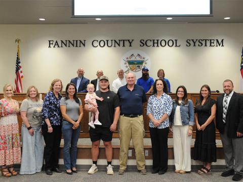 The new gym on the Fannin County High School campus will be named the Coach Johnny Farmer Gymnasium. The announcement came during the May 8 meeting of the Board of Education. Shown following the announcement are, from left, front, Dr. Connie Huff, Tara Queen, Suzianne Pass, Melissa Patton, Navy Grace held by Jordan Farmer, Farmer, Ashley H. Weeks, Shannon York, Brittney Stinnett, Dr. Scott Ramsey; and, back, Mike Cole, Bobby Bearden, Adam Born and Robert Patterson, and Shannon Dillard.