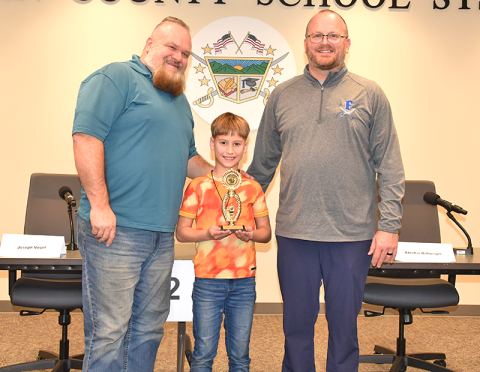 Abishai Rittweger was joined by his step dad, Charles Tucker, and East Fannin Elementary Principal Mathew Price, right, after he accepted his trophy for winning the Fannin County School District Spelling Bee last week.