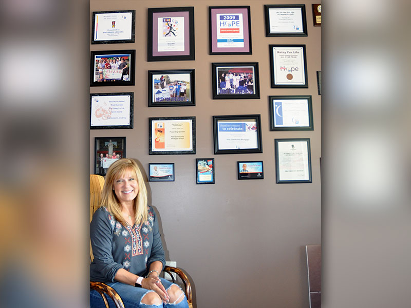 Tracie Griffith, owner and mortgage broker of First Community Mortgage, sits next to the office’s wall showcasing many of the causes the business has sponsored and supported over the years, such as Relay for Life, St. Jude and Toys for Tots.