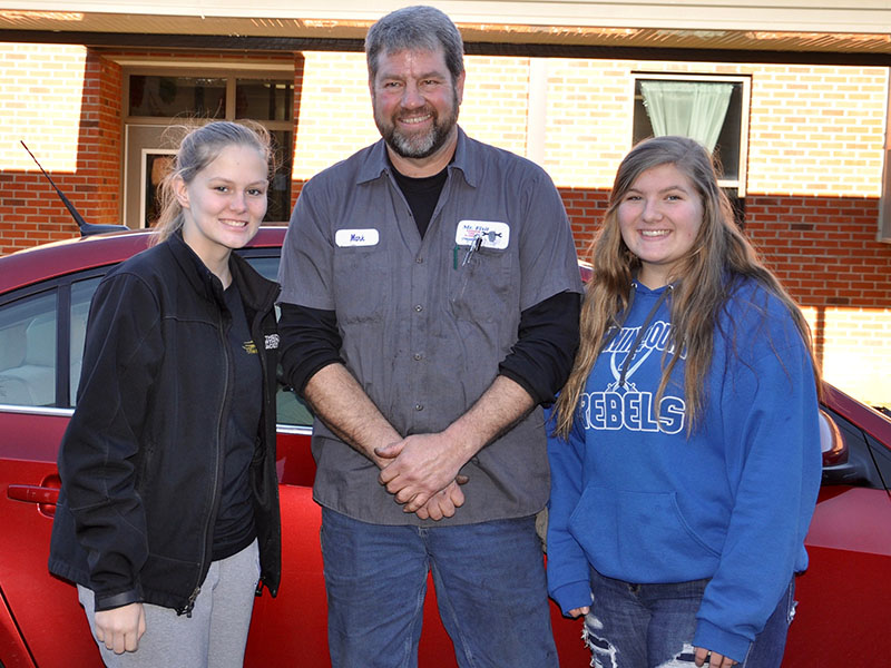 Fannin County High School (FCHS) student Alexis Hill, left, Mechanic Mark Keaton and FCHS student Bailey Pettit smile after learning to change a tire, check a tire’s air pressure and check a car’s oil Thursday, December 5.
