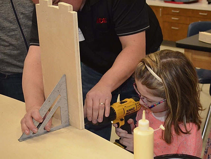 A student uses a drill with the help of a volunteer as they constructed a soap box derby car. The event re-enforces STEM lessons taught in school and also acts as a fundraiser for schools.