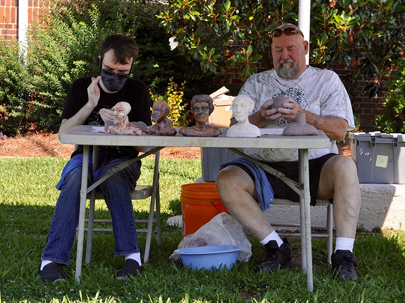 Fine Art Potters, left, Devin Weeks and Mike Lalone sculpt out some figures outside of the Crossroads: Change in Rural America exhibition at The Art Center Saturday, June 20.