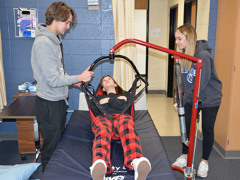 Certified Nursing Assistant (CNA) students Michael Treon and Riley Reebes demonstrate the proper way to use a life for getting a patient out of a bed using student Devan Hogsed as the patient.