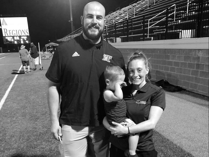 Logan Daves is pictured smiling with his wife Macy and son Laiken on the field at Toombs County High School where Daves serves as the Offensive Coordinator and assistant coach of the wrestling team. 