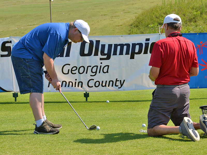 Taylor Carroll lines up a chip shot, which must go over the Special Olympics banner, as coach Craig Hartman keeps a close eye on the golfer’s expertise.
