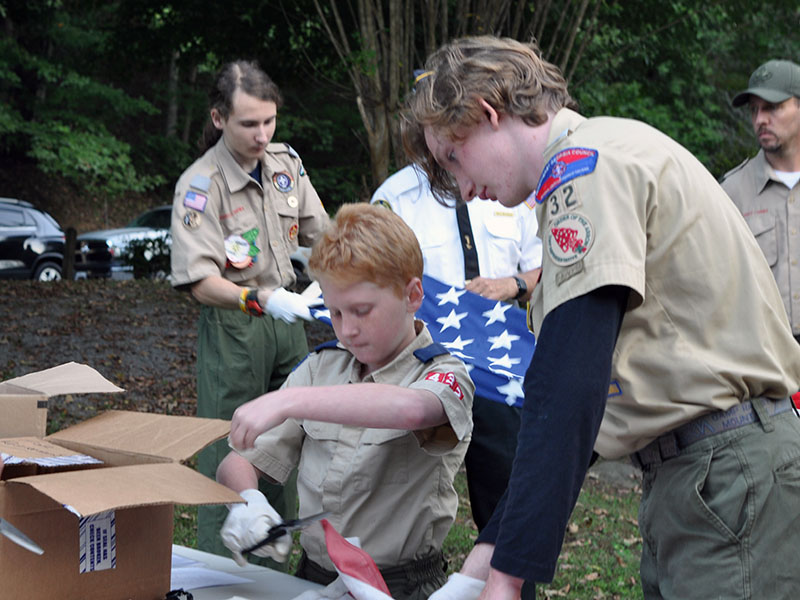 Mathew Monroe of Boy Scout Troop 32 helps a young Cub Scout, Ethan Threet, during the ceremony at Ron Henry Horseshoe Bend Park last Tuesday evening. They are working together to inspect and cut a flag separating the canton from the stripes so it can be retired properly.